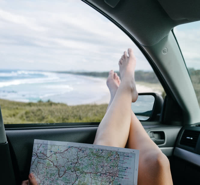 Person sitting in car and holding a map with feet out the window near the beach