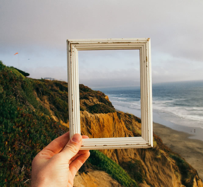 Hand holding up an empty picture frame in front of beachfront cliffs